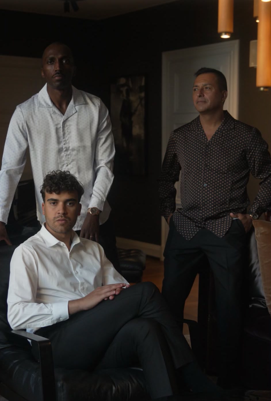 Three men of different generations at the house in the evening: one seated in a white shirt, two standing in printed shirts.